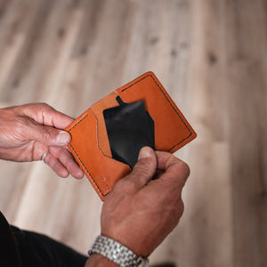A close-up of a person's hands holding an open tan leather cardholder wallet with a black card inside, set against a blurred wooden floor background.