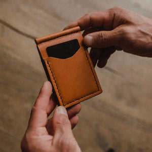 A close-up of a person's hands holding a slim, tan leather card holder wallet with a black card insert, set against a blurred wooden background. Handmade in Peterborough Ontario by jesse bateson of Solid Leather 