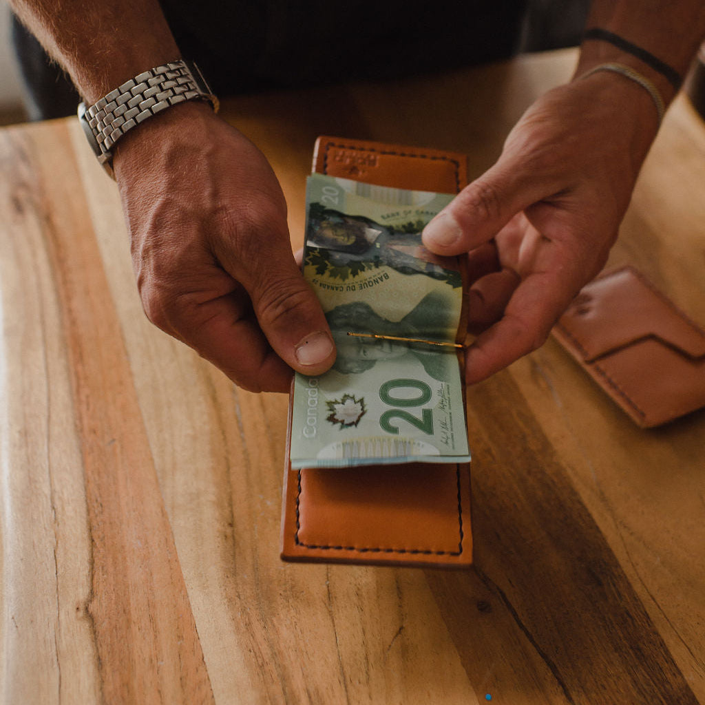 A close-up of a person holding a tan leather money clip wallet open on a wooden table, with Canadian 20 dollar notes inserted. A second closed wallet lies nearby on the table.